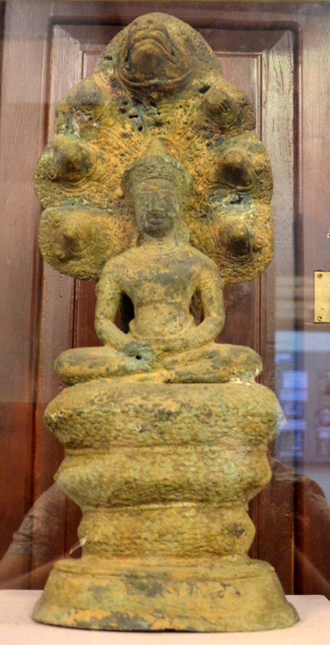 Buddha sheltered by the naga king: bronze, Ankgor period style: This sculpture represents the Buddha as both a monk and a king, with rich royal jewelry worn over simple robes. The image of the Buddha being sheltered by the seven-headed naga king Mucalinda became iconic in Angkorian times. The Buddha was meditating on the shore of the lake during the 6th week after attaining enlightenment and was caught by a raging storm. The serpent king emerged from his home in the lake and unfurled his hoods to protect the Buddha.