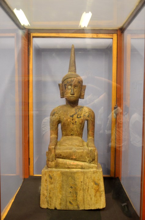 The Tai Leu Masterpiece Buddha (circa 1850) This exceptionally large teak wooden Buddha was most likely carved in the Hongsa region by a Tai Leu carver. the anatomy of the statue is advanced; note how the Buddha's gently sloping shoulders and pot-belly represent old age so directly. The carver was probably professional, possibly one of the artisans of the royal court of Luang Prabang. It was originally covered in gold leaf and traces of it and lacquer can still be seen. The statue was decorated by gem stones on the torso (note small round holes) The remaining eye is a piece of old mirror, a scarce commodity back then. The most unusual aspect is the two-hands-down pose: Buddha giving his last teaching. Not one Buddha in 100,000 is in this provocative death pose. The wooden Buddhas are carved for a blessing or to remember a loved one. Sometimes, they take on a community role. One old carver said "During the war we had nothing. We made the Buddha any way that we could." Larger Buddhas like this were made for group merit. It might have been carved as an offering during a famine or drought. One of the most amazing statues leave their habitat, their unique history is lost forever. This statue was bought in a Bangkok Junk Store for $400. Preserve Lao Culture.